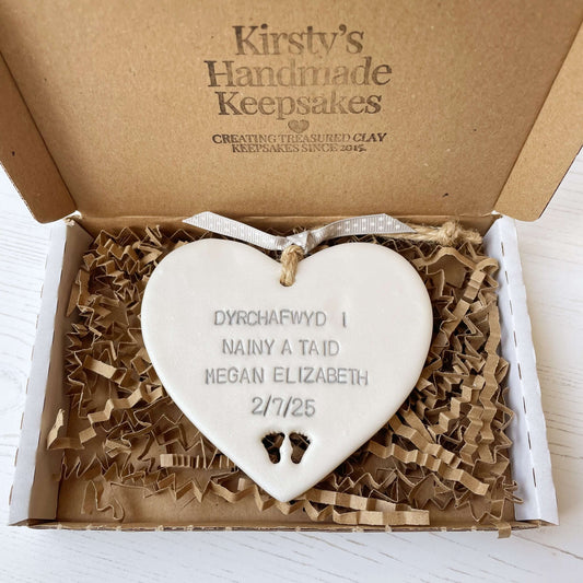 Pearlised white clay hanging heart with baby feet cut out of the bottom and grey personalisation, the heart is personalised with DYRCHAFWYD I NAINY & TAID MEGAN ELIZABETH 2/7/25