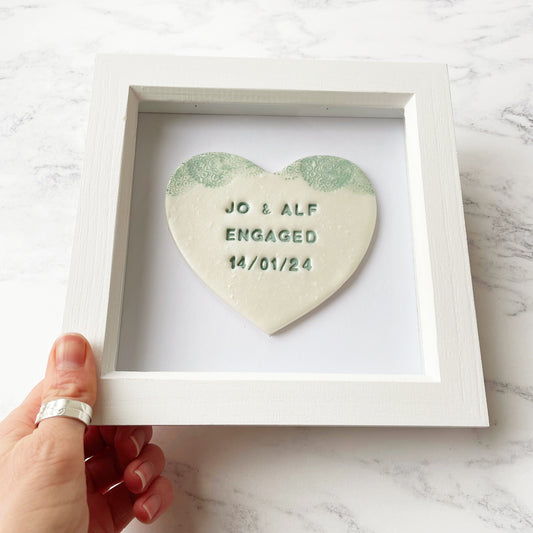 Personalised framed engagement gift, pearlised white clay heart with a white lace edge at the top of the heart in a white box frame, the heart is personalised with J0 & ALF ENGAGED 14/01/24
