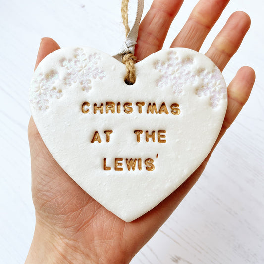 Personalised family Christmas heart ornament, pearlised white clay with CHRISTMAS AT THE LEWIS’ painted gold, decorated with 2 iridescent glitter snowflakes on either side of the top of the heart