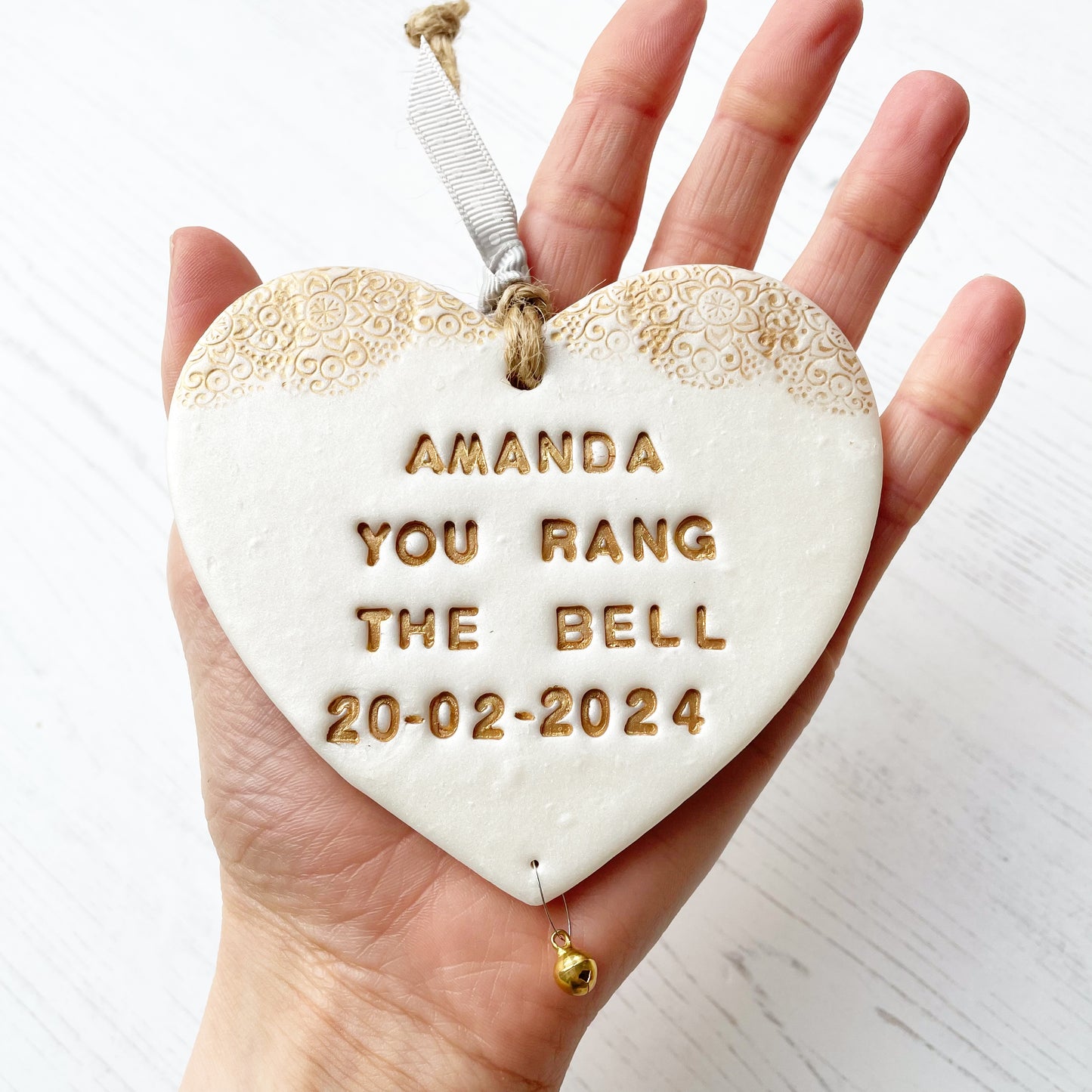 Personalised cancer survivor gift, pearlised white clay hanging heart with a gold lace edge at the top of the heart and a gold bell hanging below, the heart is personalised with AMANDA YOU RANG THE BELL 20-02-2024