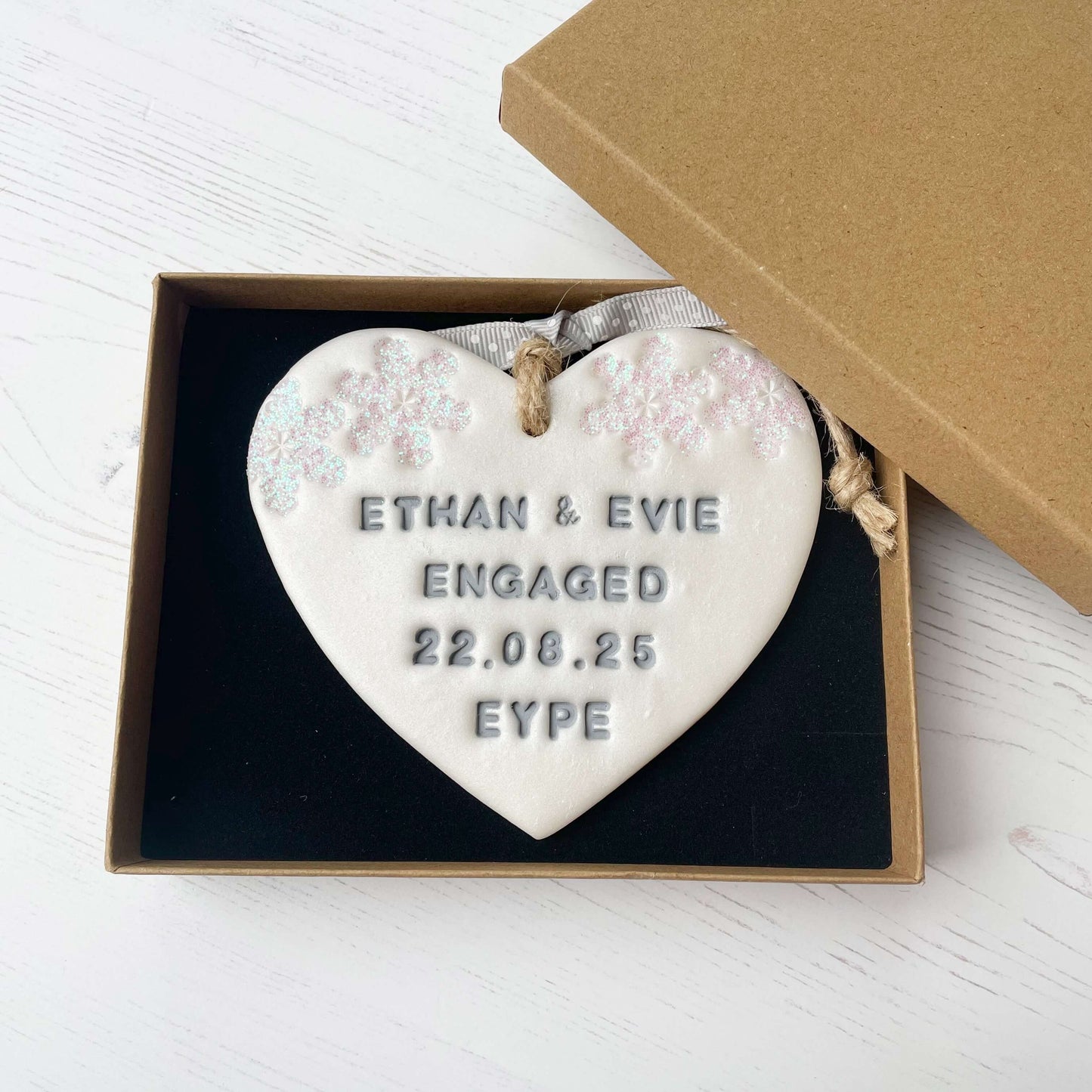Personalised first Christmas engaged heart ornament, pearlised white clay with ETHAN & EVIE ENGAGED 22.08.25 EYPE painted grey, decorated with 2 iridescent glitter snowflakes on either side of the top of the heart