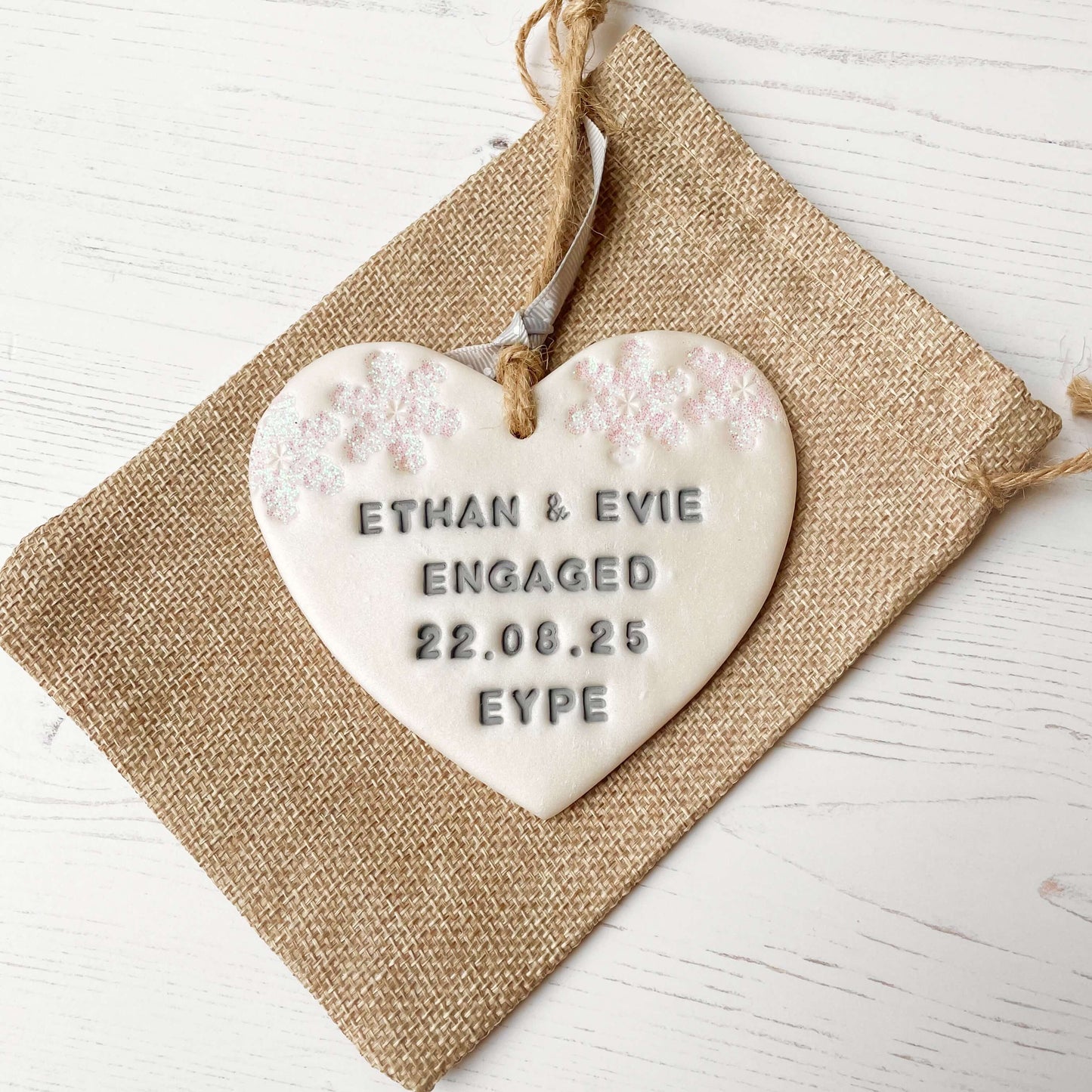 Personalised first Christmas engaged heart ornament, pearlised white clay with ETHAN & EVIE ENGAGED 22.08.25 EYPE painted grey, decorated with 2 iridescent glitter snowflakes on either side of the top of the heart