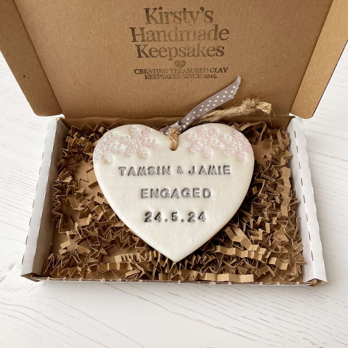 Personalised first Christmas engaged heart ornament, pearlised white clay with TAMSIN & JAMIE ENGAGED 24.5.24 painted grey, decorated with 2 iridescent glitter snowflakes on either side of the top of the heart
