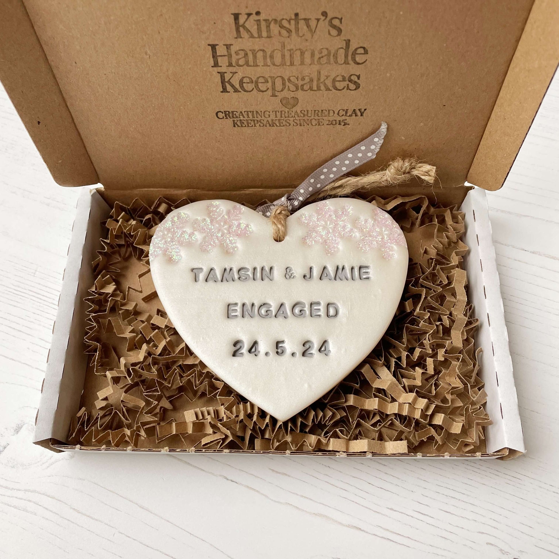Personalised first Christmas engaged heart ornament, pearlised white clay with TAMSIN & JAMIE ENGAGED 24.5.24 painted grey, decorated with 2 iridescent glitter snowflakes on either side of the top of the heart