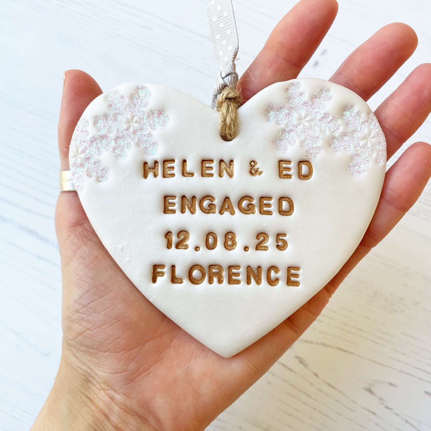 Personalised first Christmas engaged heart ornament, pearlised white clay with HELEN & ED ENGAGED 12.08.25 FLORENCE painted gold, decorated with 2 iridescent glitter snowflakes on either side of the top of the heart