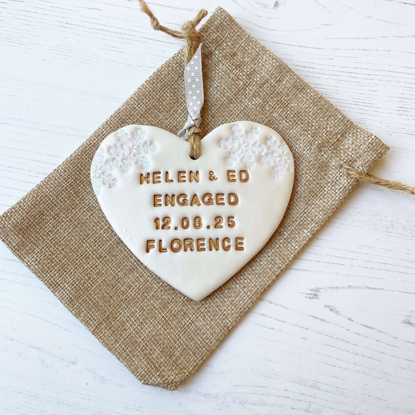 Personalised first Christmas engaged heart ornament, pearlised white clay with HELEN & ED ENGAGED 12.08.25 FLORENCE painted gold, decorated with 2 iridescent glitter snowflakes on either side of the top of the heart