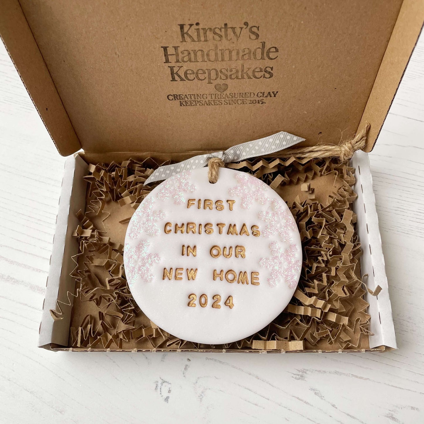 Personalised first Christmas in our new home bauble ornament, pearlised white round clay with FIRST CHRISTMAS IN OUR NEW HOME 2024 in gold, decorated with 3 iridescent glitter snowflakes on either side of the bauble
