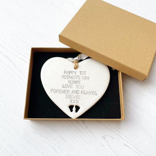Personalised first Mother’s Day gift, pearlised white hanging clay heart with baby feet cut out at the bottom of the heart, the heart is personalised in grey with HAPPY 1ST MOTHER'S DAY MOMMY, LOVE YOU FOREVER AND ALWAYS, STEVIE XXX In a luxury Kraft brown gift box