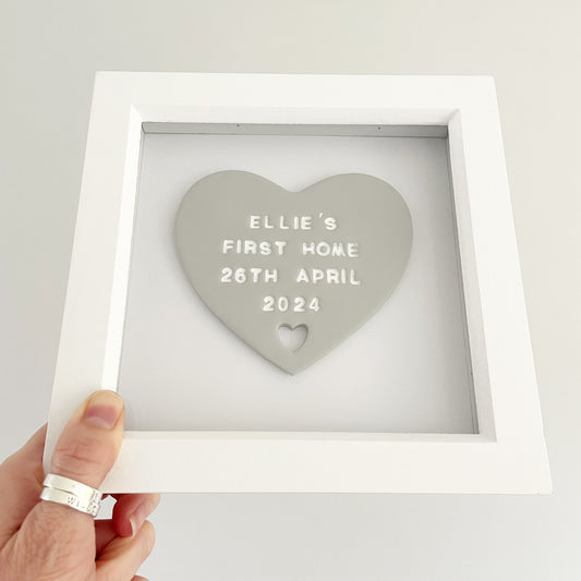 Personalised housewarming new home gift, grey clay heart with a heart cut out at the bottom in a white box frame, the heart is personalised in white with ELLIE’S FIRST HOME 26TH APRIL 2024