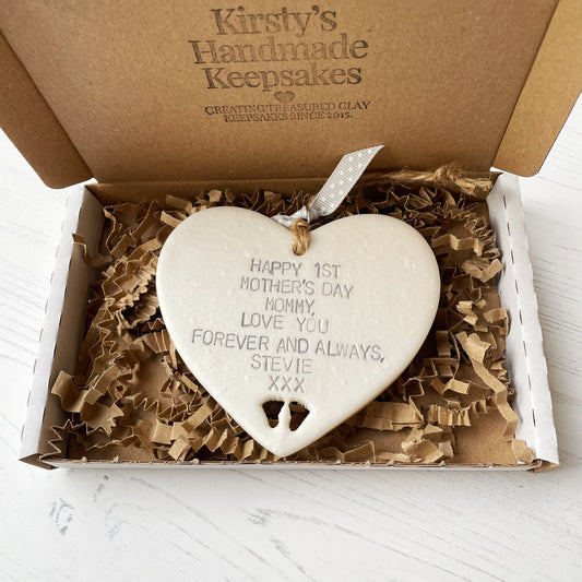 Personalised first Mother’s Day gift, pearlised white hanging clay heart with baby feet cut out at the bottom of the heart, the heart is personalised in grey with HAPPY 1ST MOTHER'S DAY MOMMY, LOVE YOU FOREVER AND ALWAYS, STEVIE XXX In a postal box with brown Kraft shredded zigzag paper