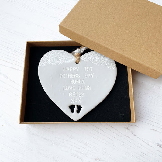 Personalised first Mother’s Day keepsake gift, grey clay hanging heart with baby feet cut out at the bottom of the heart and a white lace edge at the top of the heart, the heart is personalised with HAPPY 1ST MOTHER'S DAY MUMMY, LOVE FROM BETSY XXX in a brown Kraft luxury gift box