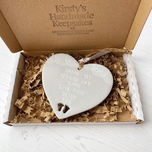 Personalised first Mother’s Day keepsake gift, grey clay hanging heart with baby feet cut out at the bottom of the heart and a white lace edge at the top of the heart, the heart is personalised with HAPPY 1ST MOTHER'S DAY MUMMY, LOVE FROM BETSY XXX in a postal box with brown Kraft shredded zigzag paper