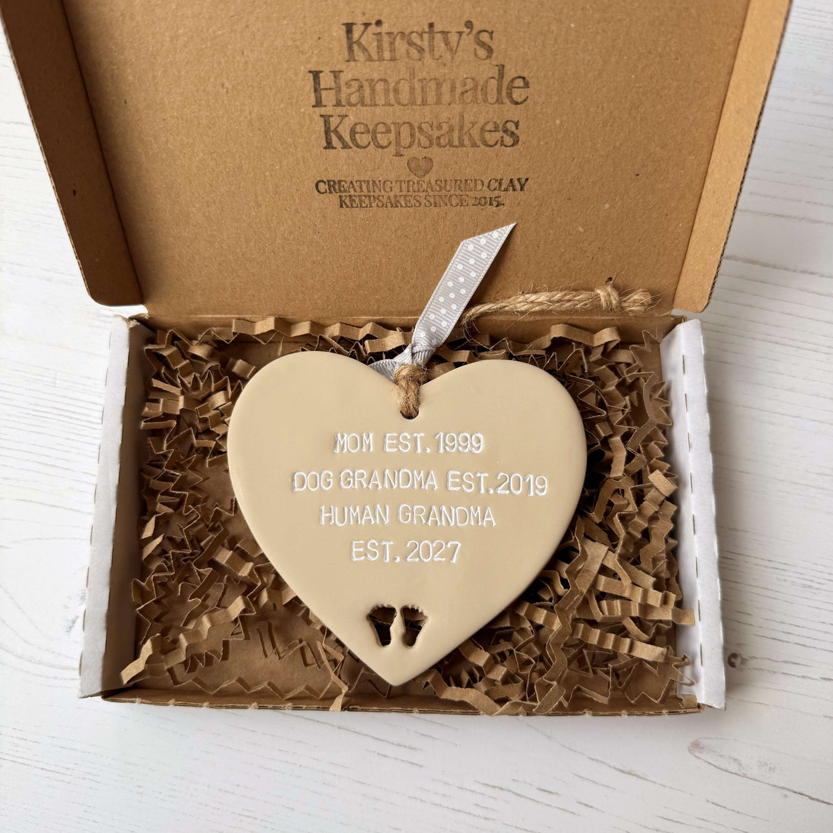 Beige clay hanging heart with baby feet cut out of the bottom and white personalisation, the heart is personalised with MOM EST. 1999 DOG GRANDMA EST. 2019 HUMAN GRANDMA EST. 2027