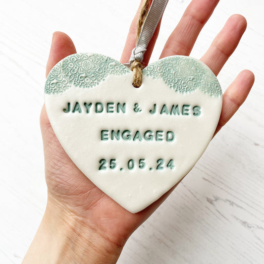 Personalised engagement gift, pearlised white clay heart with a sage green lace edge at the top of the heart with jute twine for hanging, the heart is personalised with JAYDEN & JAMES ENGAGED 25.05.24