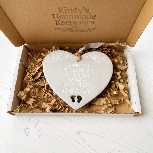 Personalised Godfather gift, grey clay hanging heart with baby feet cut out of the bottom and white lace edge at the top of the heart, the heart is personalised with THANK YOU FOR BEING MY GODFATHER NATHAN. LOVE WREN 23/06/2024 in a postal box with brown Kraft shredded zigzag paper
