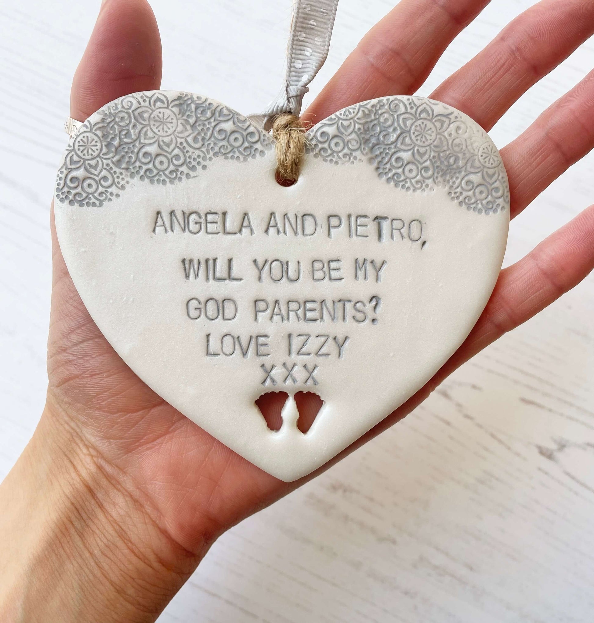Will you be my godparents gift pearlised white clay hanging heart with baby feet cut out of the bottom grey lace edge at the top the heart is personalised with ANGELA & PIETRO, WILL YOU BE MY GODPARENTS? LOVE IZZY XXX