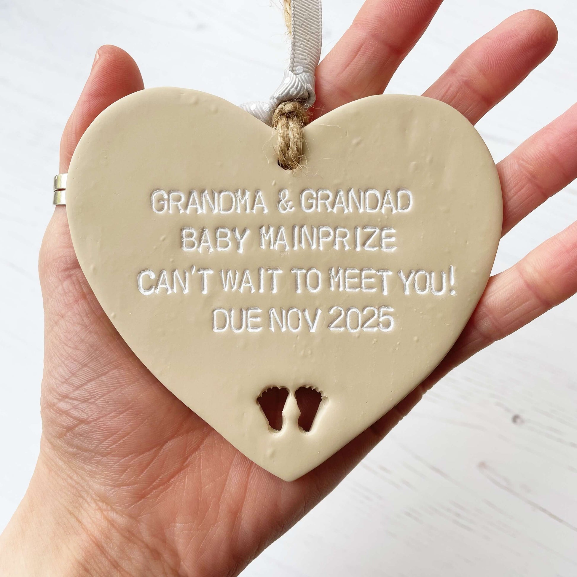 Beige clay hanging heart with baby feet cut out of the bottom and white personalisation, the heart is personalised with GRANDMA AND GRANDAD BABY MAINPRIZE CAN’T WAIT TO MEET YOU DUE NOV 2025