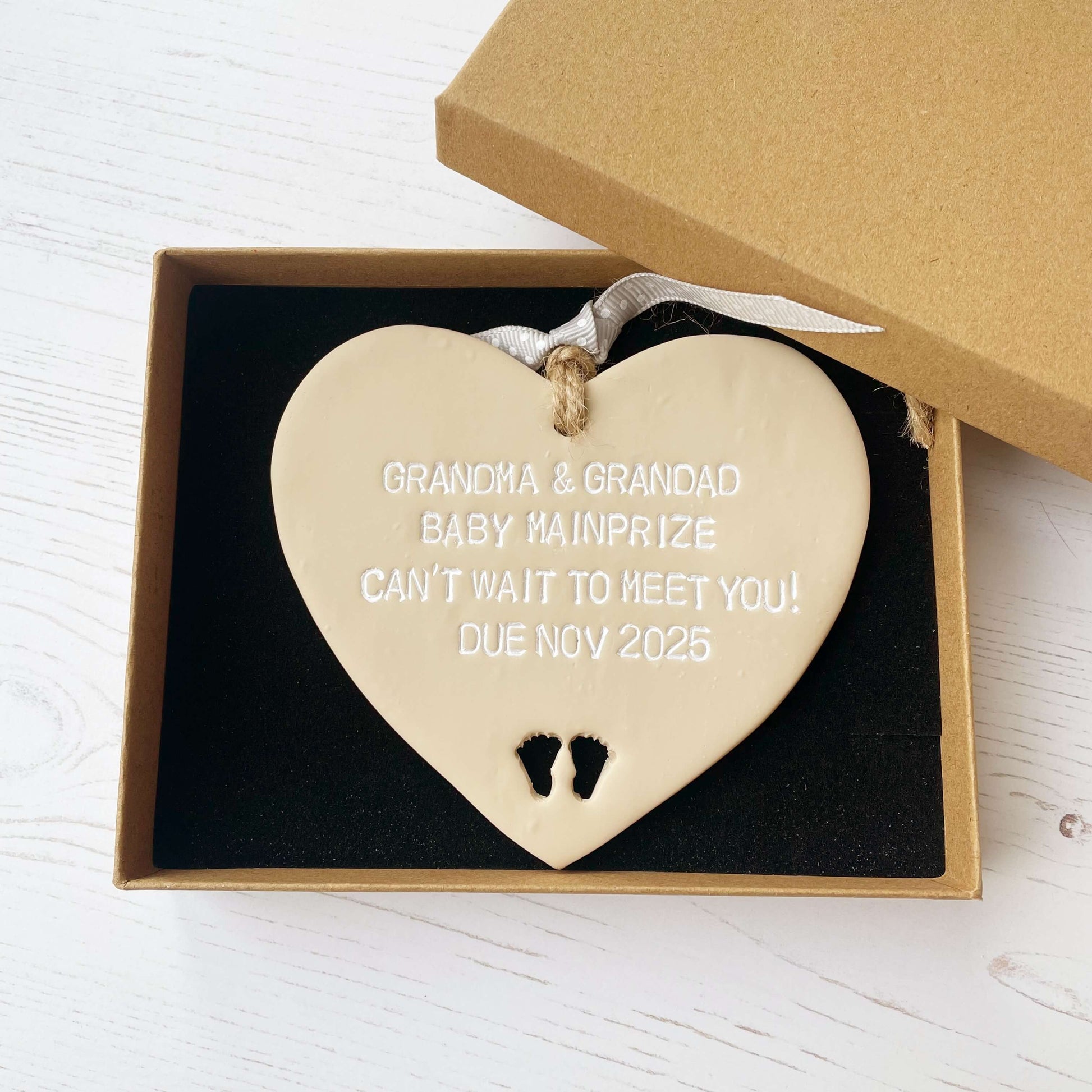 Beige clay hanging heart with baby feet cut out of the bottom and white personalisation, the heart is personalised with GRANDMA AND GRANDAD BABY MAINPRIZE CAN’T WAIT TO MEET YOU DUE NOV 2025