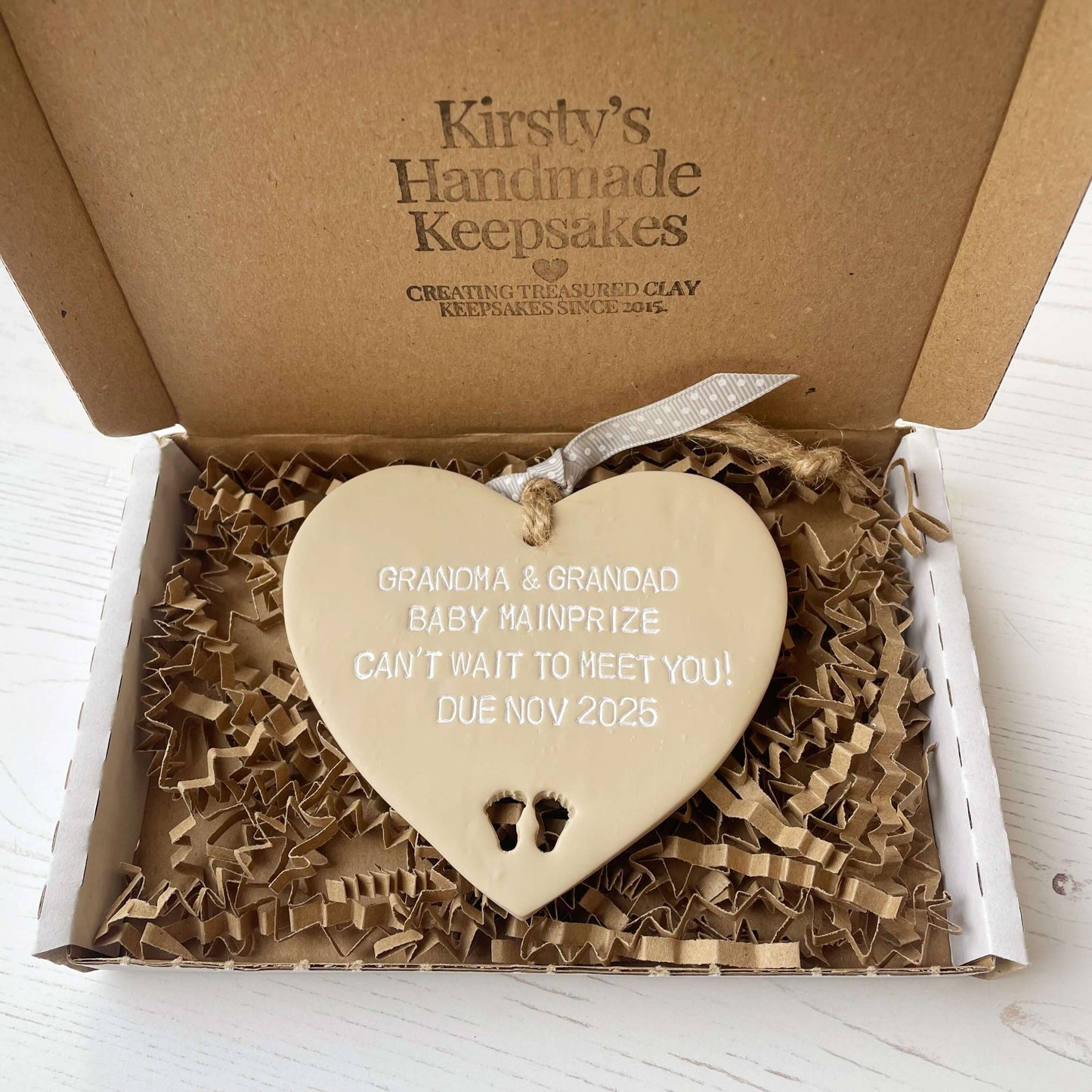 Beige clay hanging heart with baby feet cut out of the bottom and white personalisation, the heart is personalised with GRANDMA AND GRANDAD BABY MAINPRIZE CAN’T WAIT TO MEET YOU DUE NOV 2025