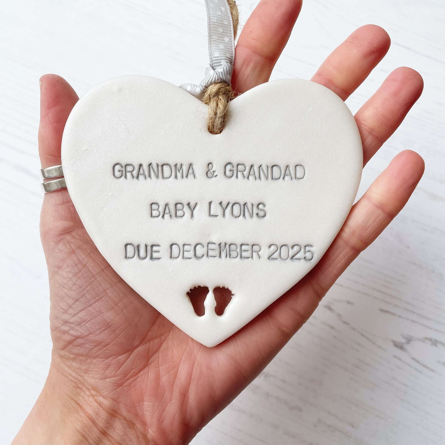 Pearlised white clay hanging heart with baby feet cut out of the bottom and grey personalisation, the heart is personalised with GRANDMA & GRANDAD BABY LYONS DUE DECEMBER 2025