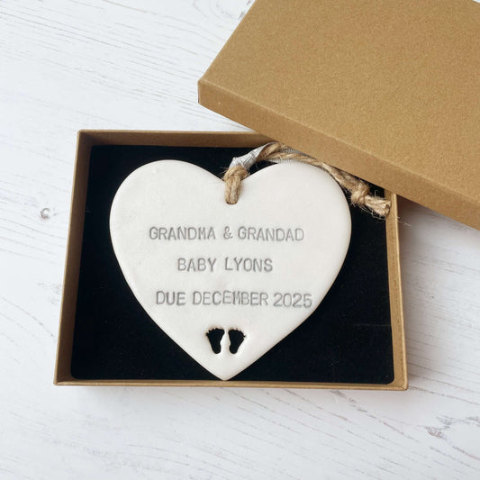 Pearlised white clay hanging heart with baby feet cut out of the bottom and grey personalisation, the heart is personalised with GRANDMA & GRANDAD BABY LYONS DUE DECEMBER 2025