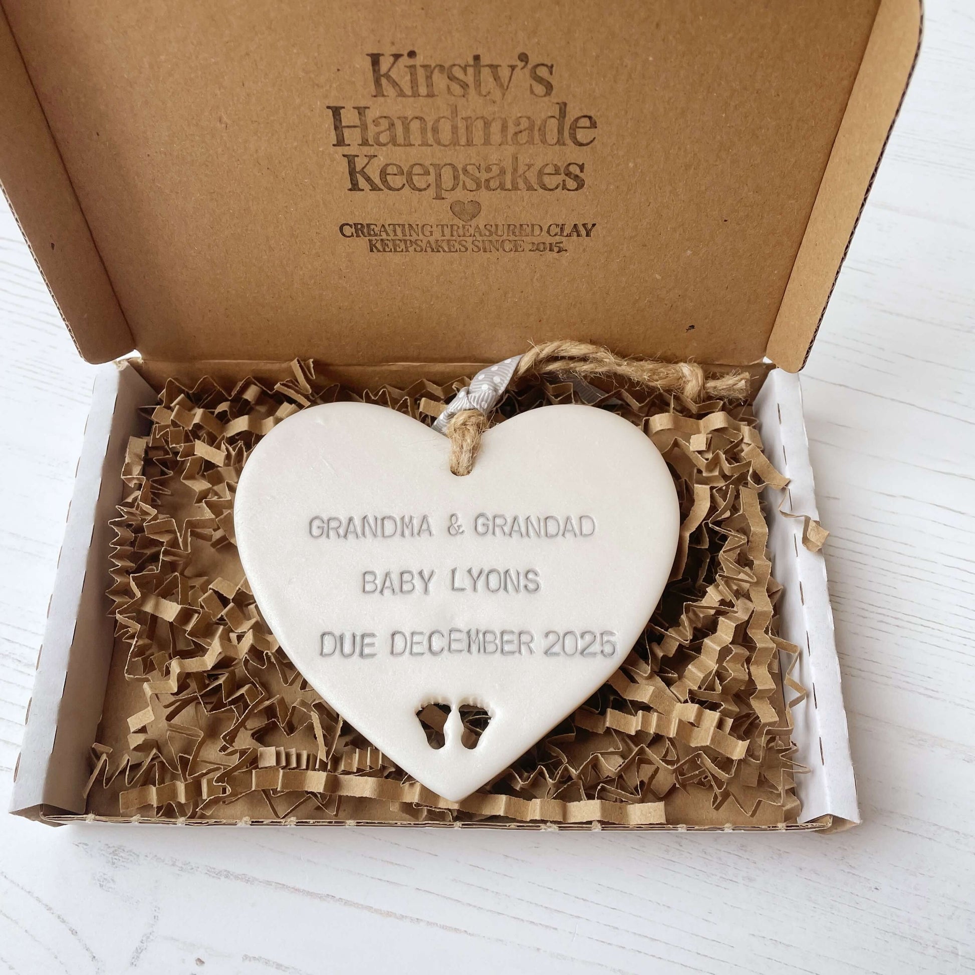 Pearlised white clay hanging heart with baby feet cut out of the bottom and grey personalisation, the heart is personalised with GRANDMA & GRANDAD BABY LYONS DUE DECEMBER 2025