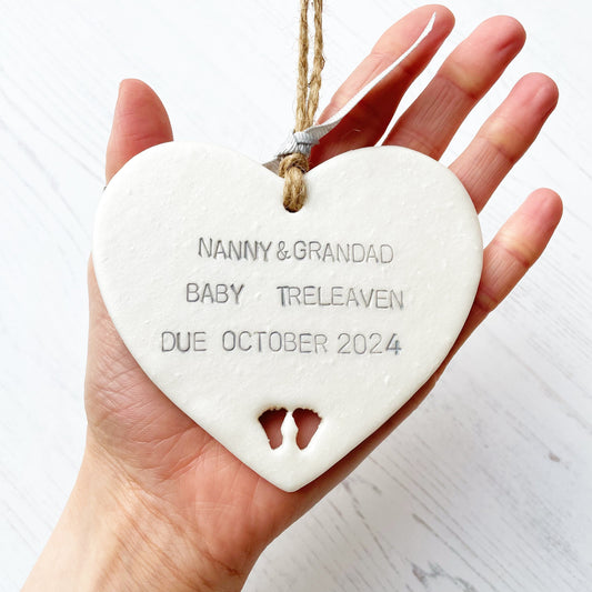 Pearlised white clay hanging heart with baby feet cut out of the bottom and grey personalisation, the heart is personalised with NANNY & GRANDAD BABY TRELEAVEN DUE OCTOBER 2024
