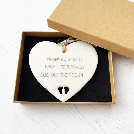 Pearlised white clay hanging heart with baby feet cut out of the bottom and grey personalisation, the heart is personalised with NANNY & GRANDAD BABY TRELEAVEN DUE OCTOBER 2024 In a brown Kraft luxury gift box