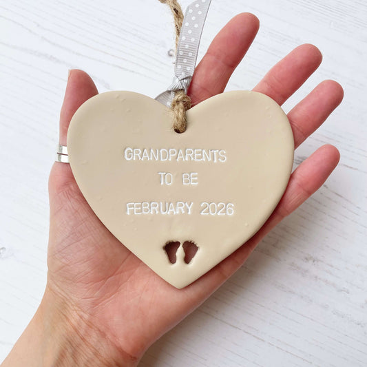 Beige clay hanging heart with baby feet cut out of the bottom and white personalisation, the heart is personalised with GRANDPARENTS TO BE FEBRUARY 2026