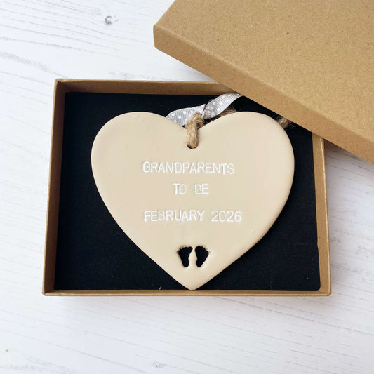 Beige clay hanging heart with baby feet cut out of the bottom and white personalisation, the heart is personalised with GRANDPARENTS TO BE FEBRUARY 2026