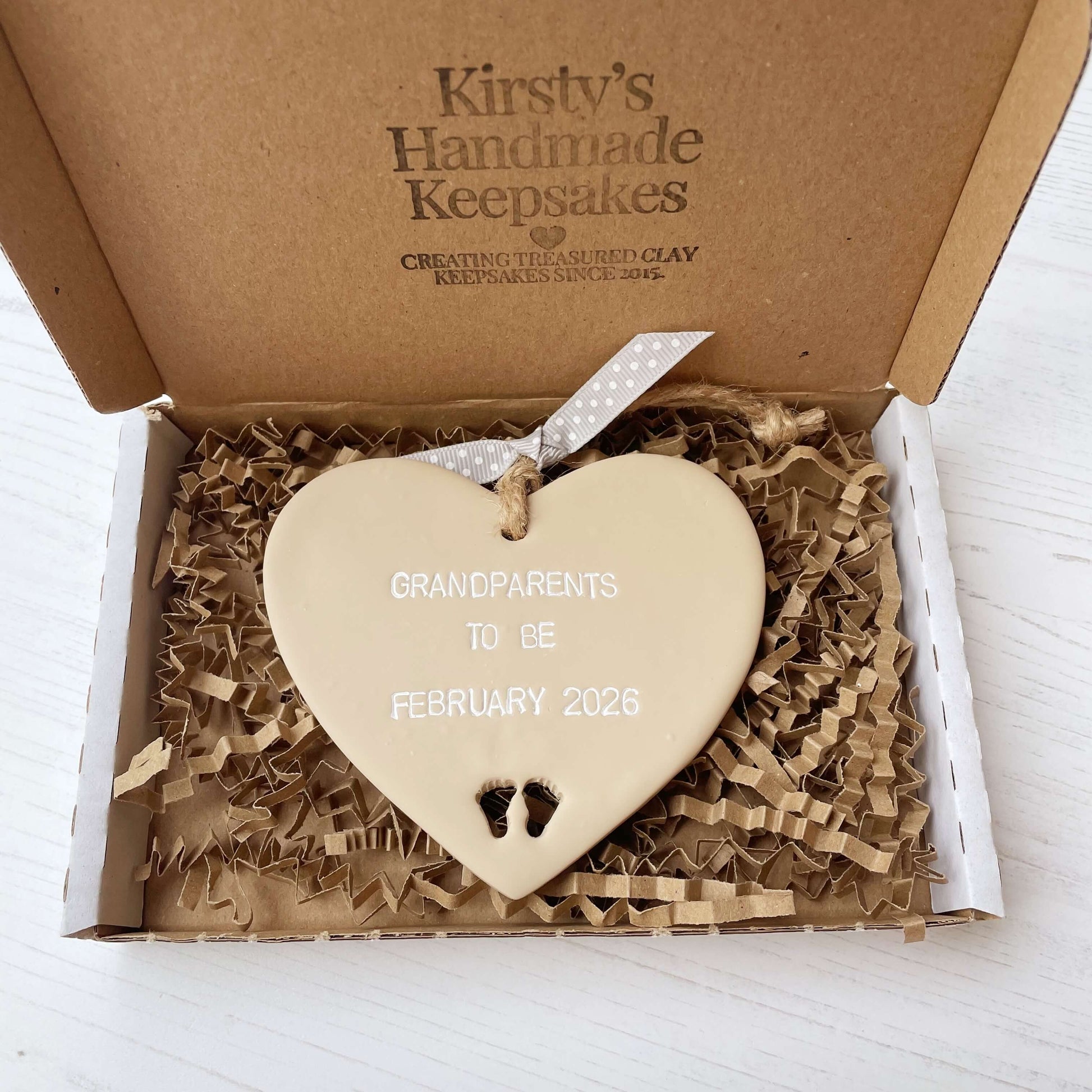 Beige clay hanging heart with baby feet cut out of the bottom and white personalisation, the heart is personalised with GRANDPARENTS TO BE FEBRUARY 2026