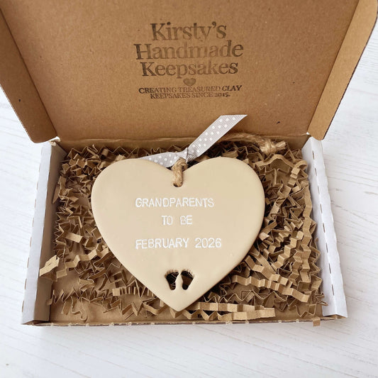 Beige clay hanging heart with baby feet cut out of the bottom and white personalisation, the heart is personalised with GRANDPARENTS TO BE FEBRUARY 2026