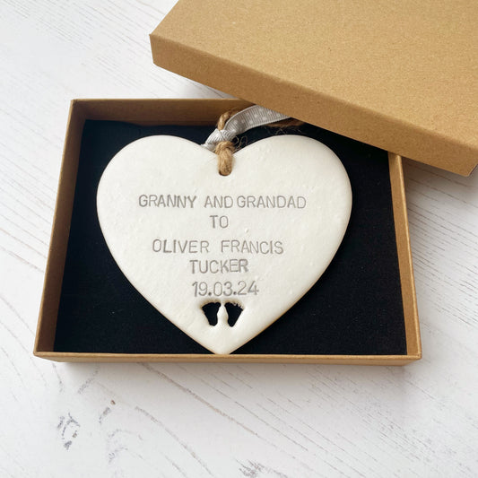 Pearlised white clay hanging heart with baby feet cut out of the bottom and grey personalisation, the heart is personalised with GRANNY AND GRANDAD TO OLIVER FRANCIS TUCKER 19.03.24 In a brown Kraft luxury gift box