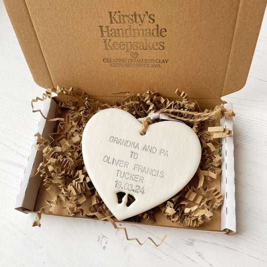 Pearlised white clay hanging heart with baby feet cut out of the bottom and grey personalisation, the heart is personalised with GRANDMA AND PA TO OLIVER FRANCIS TUCKER 19.03.24 In a postal box with brown Kraft shredded zigzag paper
