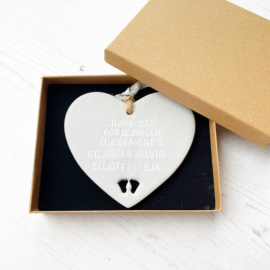 Personalised Guide parent gift, grey clay hanging heart with baby feet cut out of the bottom, the heart is personalised with THANK-YOU FOR BEING OUR GUIDEPARENT’S STEPHEN & KELVIN. ELLIOTT & EMILIA In a brown Kraft luxury gift box