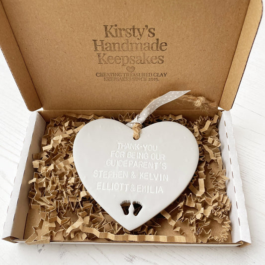 Personalised Guide parent gift, grey clay hanging heart with baby feet cut out of the bottom, the heart is personalised with THANK-YOU FOR BEING OUR GUIDEPARENT’S STEPHEN & KELVIN. ELLIOTT & EMILIA In a postal box with brown Kraft shredded zigzag paper