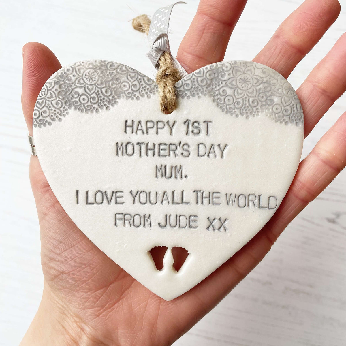 Personalised first Mother’s Day gift, pearlised white clay hanging heart with baby feet cut out at the bottom of the heart and a grey lace edge at the top of the heart, the heart is personalised with HAPPY 1ST MOTHER’S DAY MUM. I LOVE YOU ALL THE WORLD FROM JUDE XX