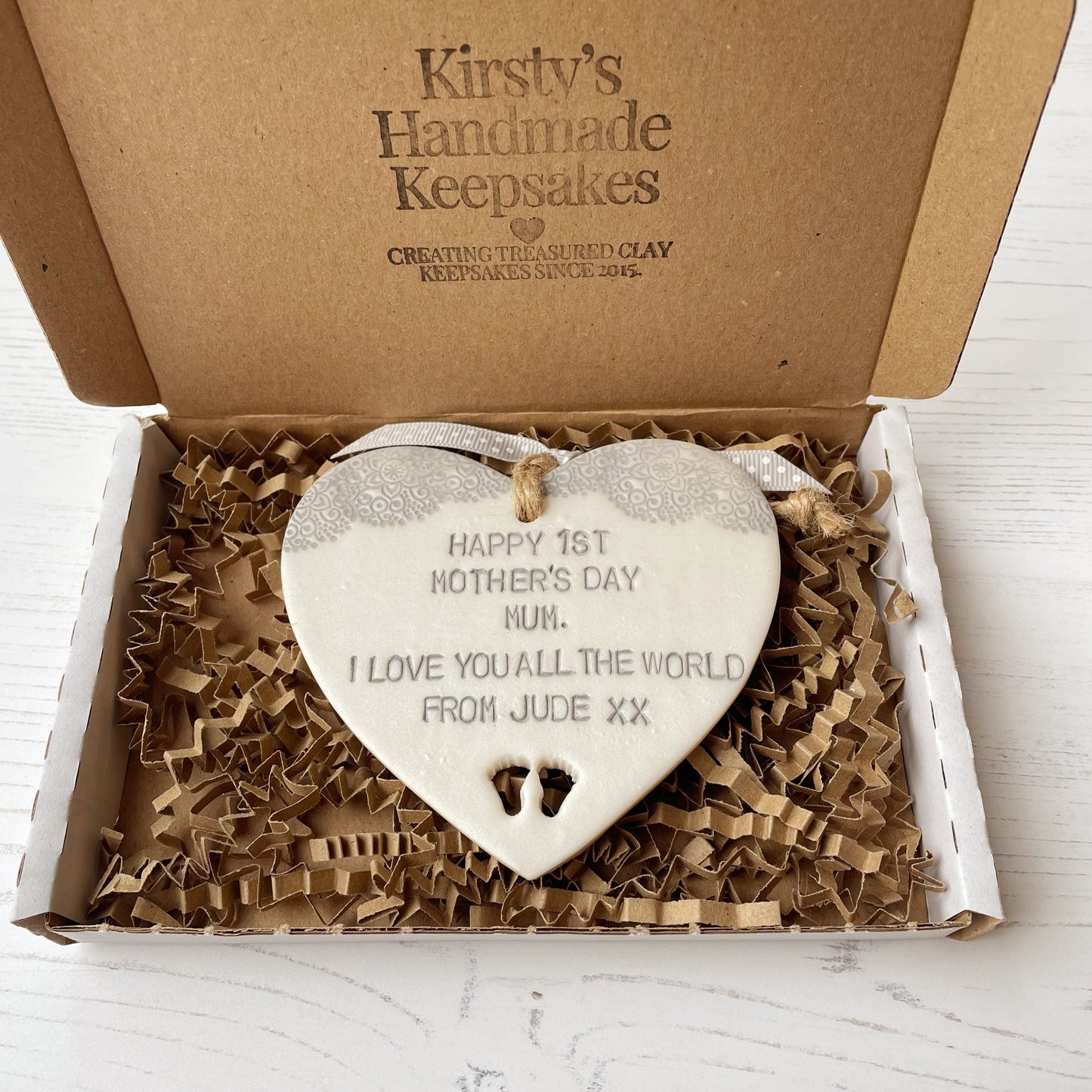 Personalised first Mother’s Day gift, pearlised white clay hanging heart with baby feet cut out at the bottom of the heart and a grey lace edge at the top of the heart, the heart is personalised with HAPPY 1ST MOTHER’S DAY MUM. I LOVE YOU ALL THE WORLD FROM JUDE XX