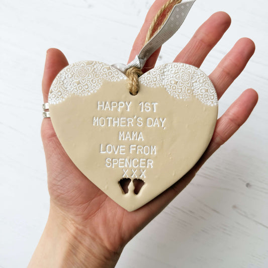 Personalised first Mother’s Day gift, beige clay hanging heart with baby feet cut out at the bottom of the heart and a white lace edge at the top of the heart, the heart is personalised with HAPPY 1ST MOTHER’S DAY MAMA, LOVE FROM SPENCER XXX