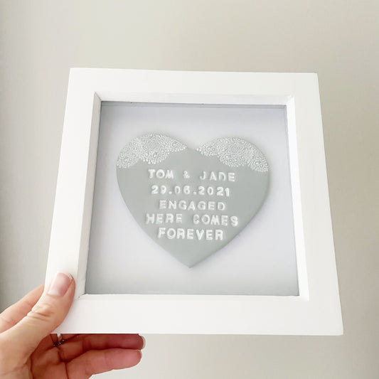 Personalised framed engagement gift, grey clay heart with a white lace edge at the top of the heart in a white box frame, the heart is personalised with TOM & JADE 29.06.2021 ENGAGED HERE COMES FOREVER