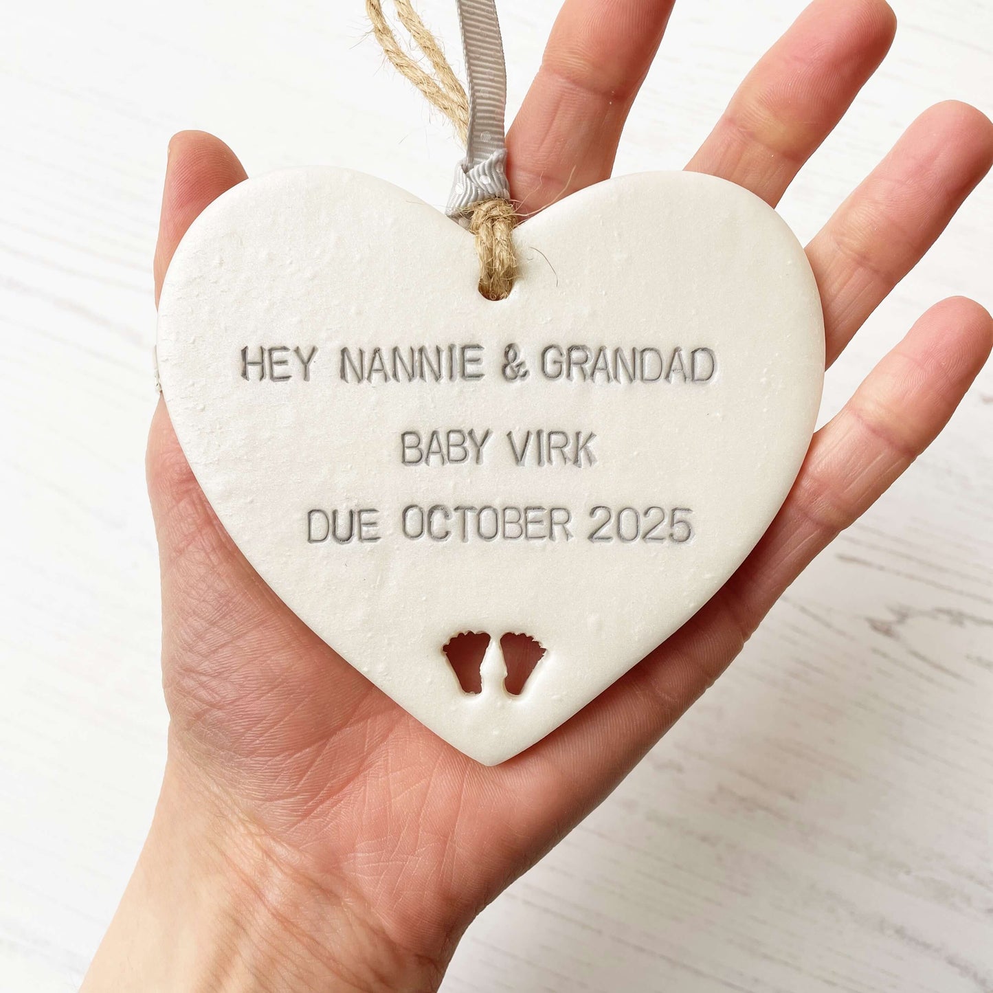 Pearlised white clay hanging heart with baby feet cut out of the bottom and grey personalisation, the heart is personalised with HEY NANNIE & GRANDAD BABY VIRK DUE OCTOBER 2025