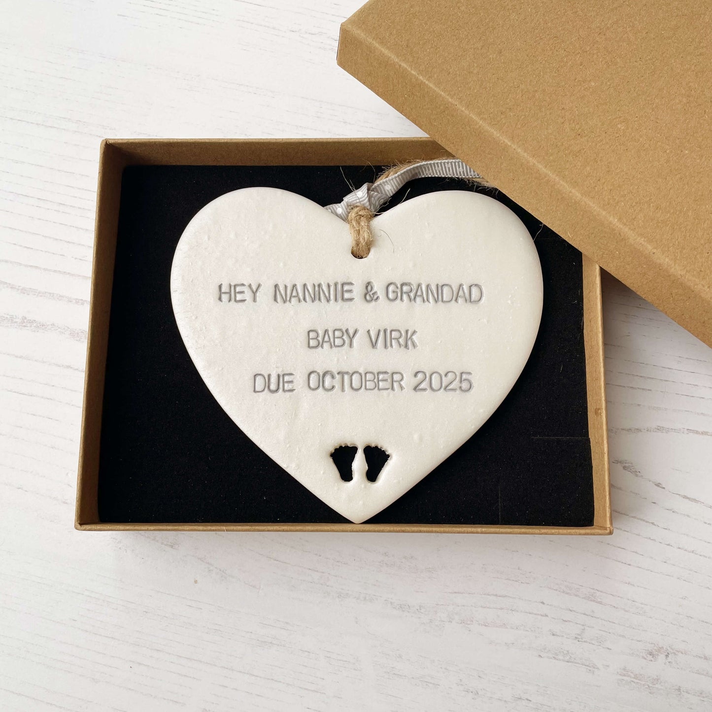 Pearlised white clay hanging heart with baby feet cut out of the bottom and grey personalisation, the heart is personalised with HEY NANNIE & GRANDAD BABY VIRK DUE OCTOBER 2025