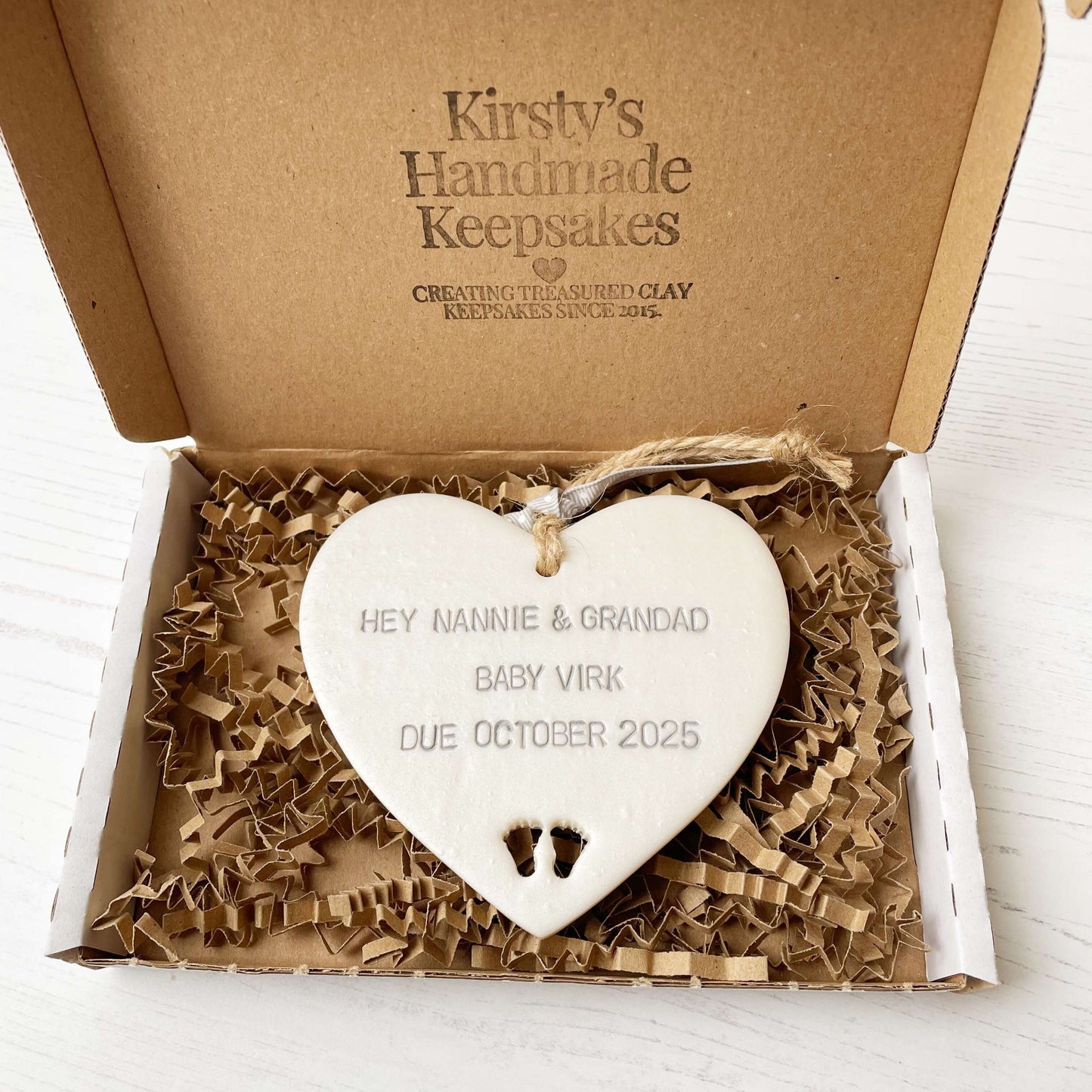 Pearlised white clay hanging heart with baby feet cut out of the bottom and grey personalisation, the heart is personalised with HEY NANNIE & GRANDAD BABY VIRK DUE OCTOBER 2025