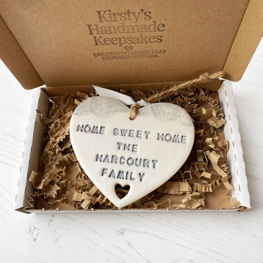 Personalised new home gift pearlised white clay heart with a grey lace edge at the top & a heart cut out at the bottom with twine for hanging personalised with HOME SWEET HOME THE HARCOURT FAMILY in a postal box with brown kraft shredded zigzag paper
