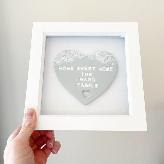 Personalised housewarming new home gift, grey clay heart with a white lace edge at the top of the heart and a heart cut out at the bottom in a white box frame, the heart is personalised with HOME SWEET HOME THE WARD FAMILY
