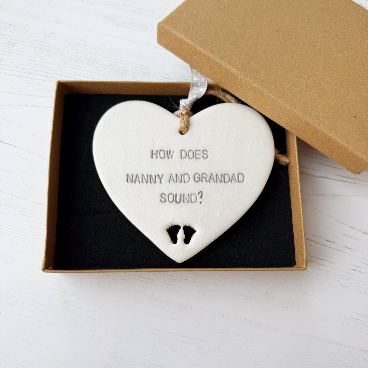 Pearlised white clay hanging heart with baby feet cut out of the bottom and grey personalisation, the heart is personalised with HOW DOES NANNY AND GRANDAD SOUND?
