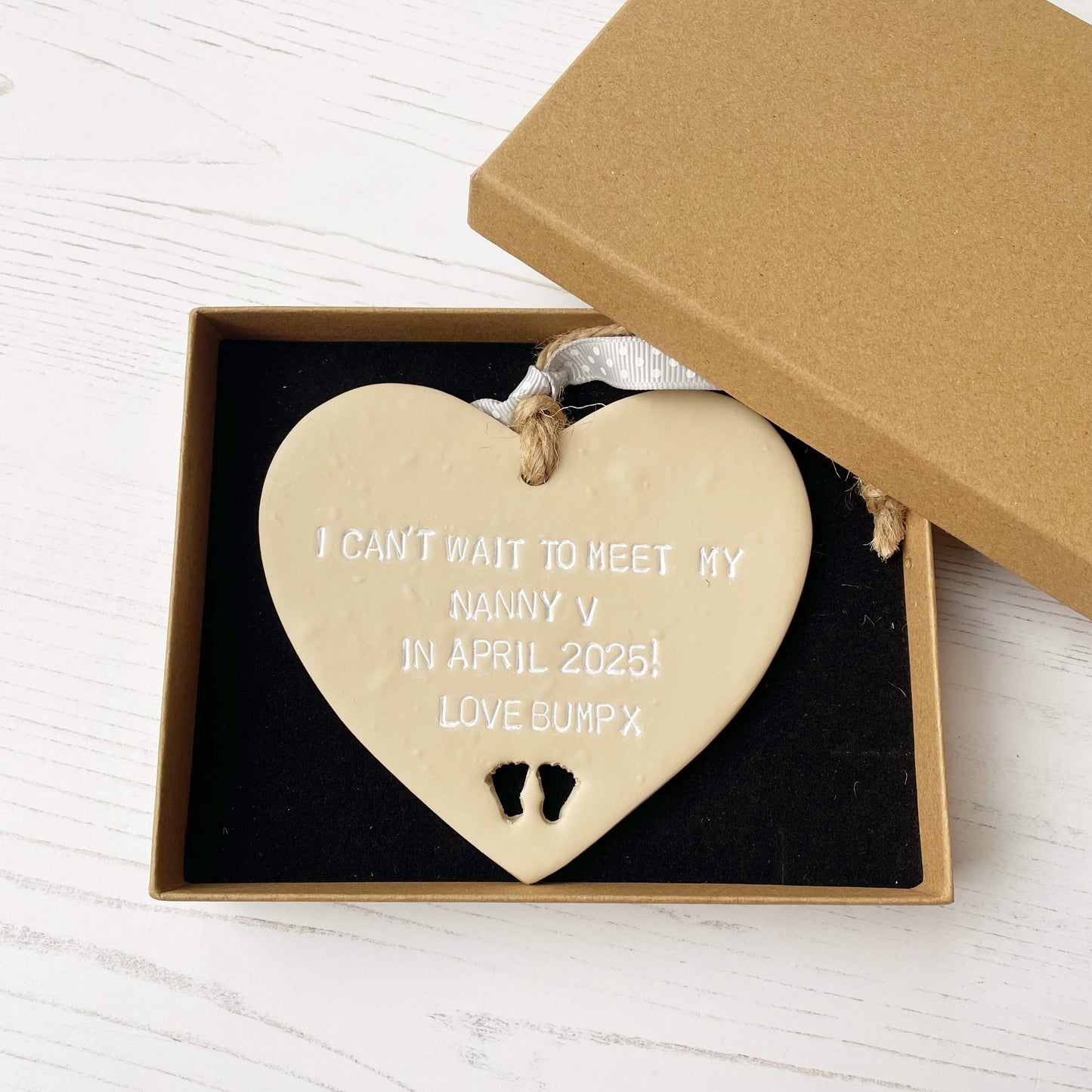 Beige clay hanging heart with baby feet cut out of the bottom and white personalisation, the heart is personalised with I CAN’T WAIT TO MEET MY NANNY V IN APRIL 2025! LOVE BUMP X
