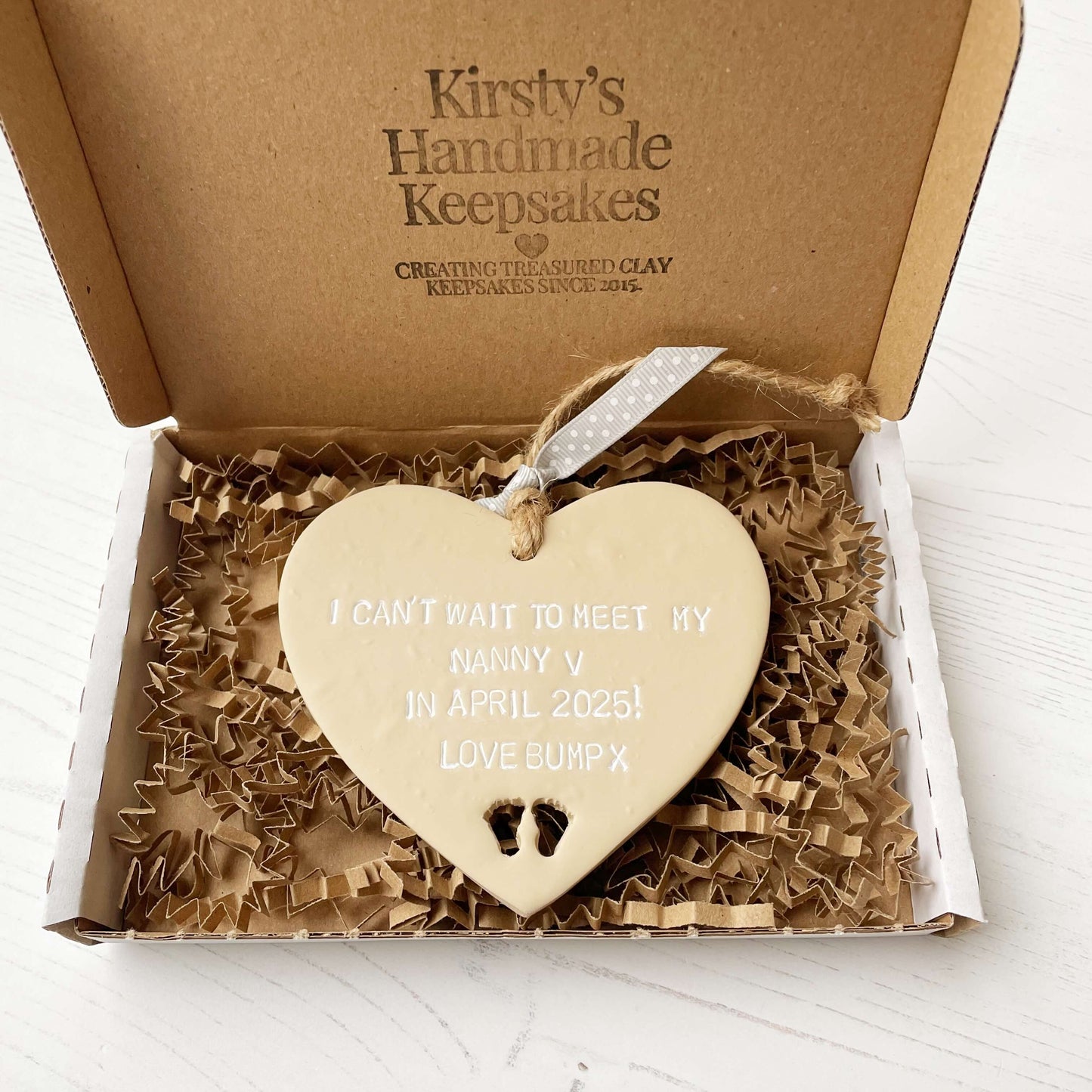 Beige clay hanging heart with baby feet cut out of the bottom and white personalisation, the heart is personalised with I CAN’T WAIT TO MEET MY NANNY V IN APRIL 2025! LOVE BUMP X