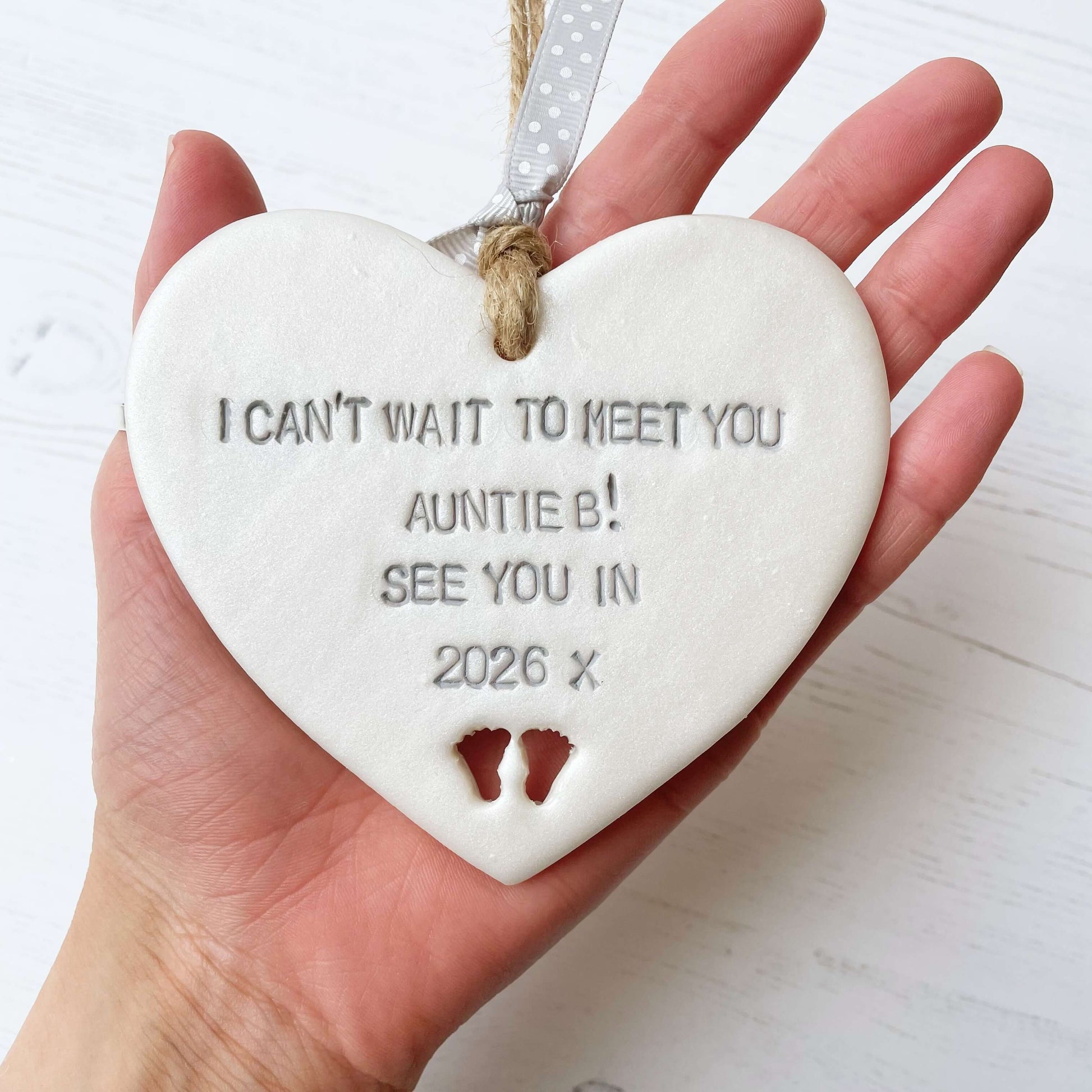 Pearlised white clay hanging heart with baby feet cut out of the bottom and grey personalisation, the heart is personalised with I CAN’T WAIT TO MEET YOU AUNTIE B! SEE YOU IN 2026 X