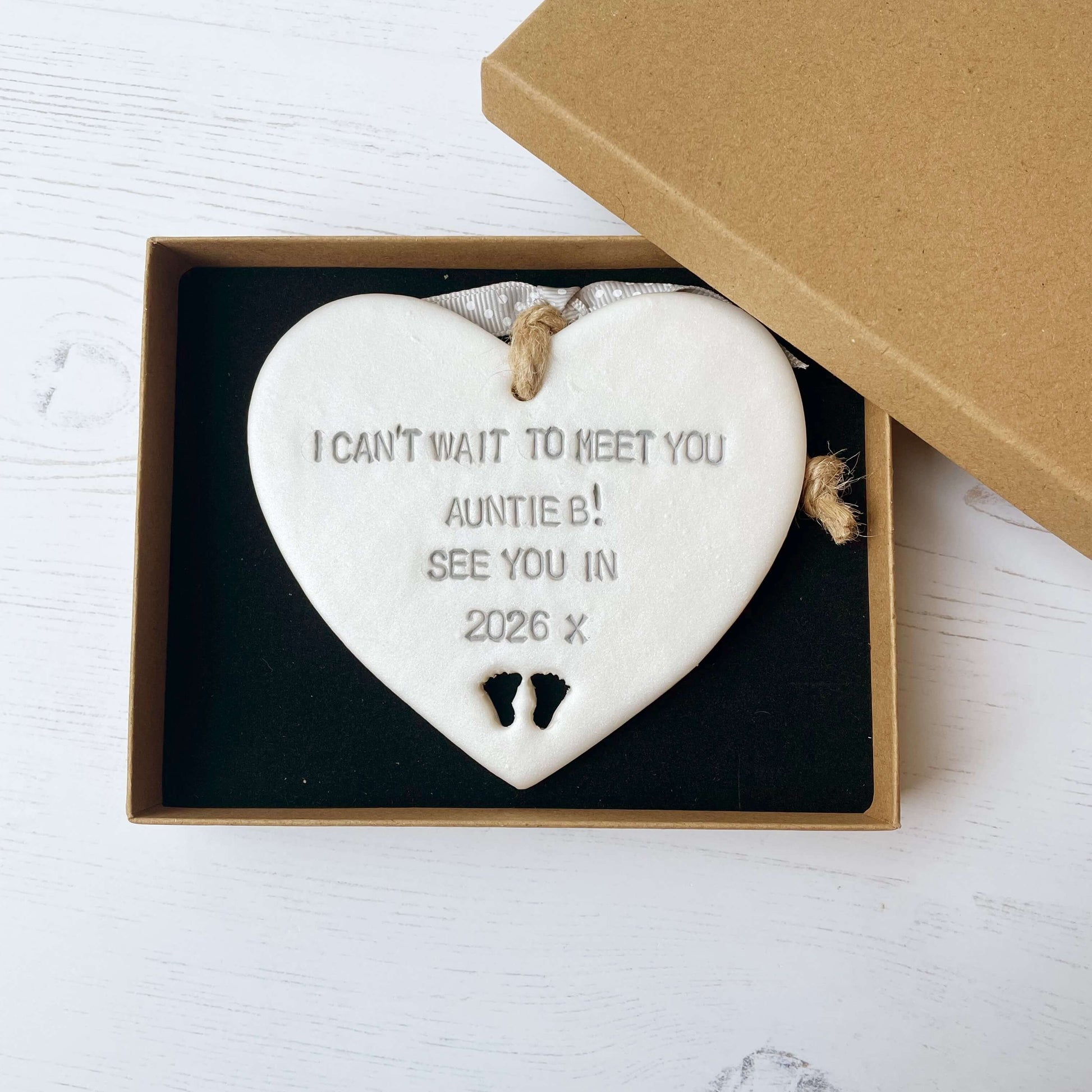Pearlised white clay hanging heart with baby feet cut out of the bottom and grey personalisation, the heart is personalised with I CAN’T WAIT TO MEET YOU AUNTIE B! SEE YOU IN 2026 X